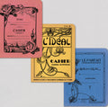 Three vintage-style notebook covers arranged overlapping on a light gray background: a pink cover with black rose illustration and text reading 'ECOLE' and 'CAHIER SPECIA', a yellow cover with ornate floral artwork and 'L'IDEAL CAHIER', and a blue cover with floral line art and 'LE PARFAIT CAHIER'.