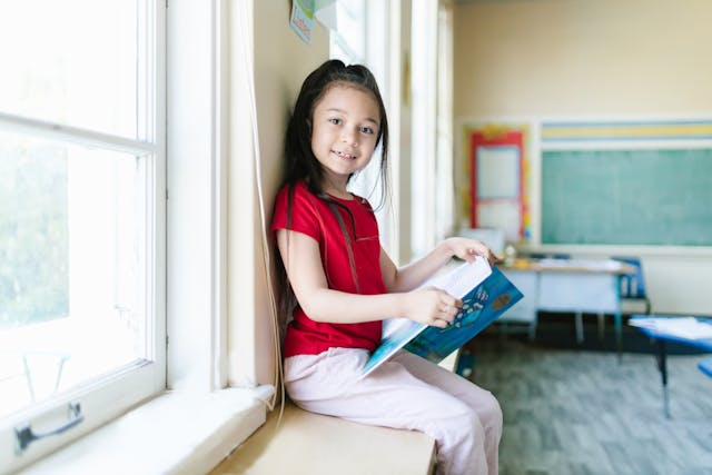 Young girl reading a book on a windowsill in a classroom
